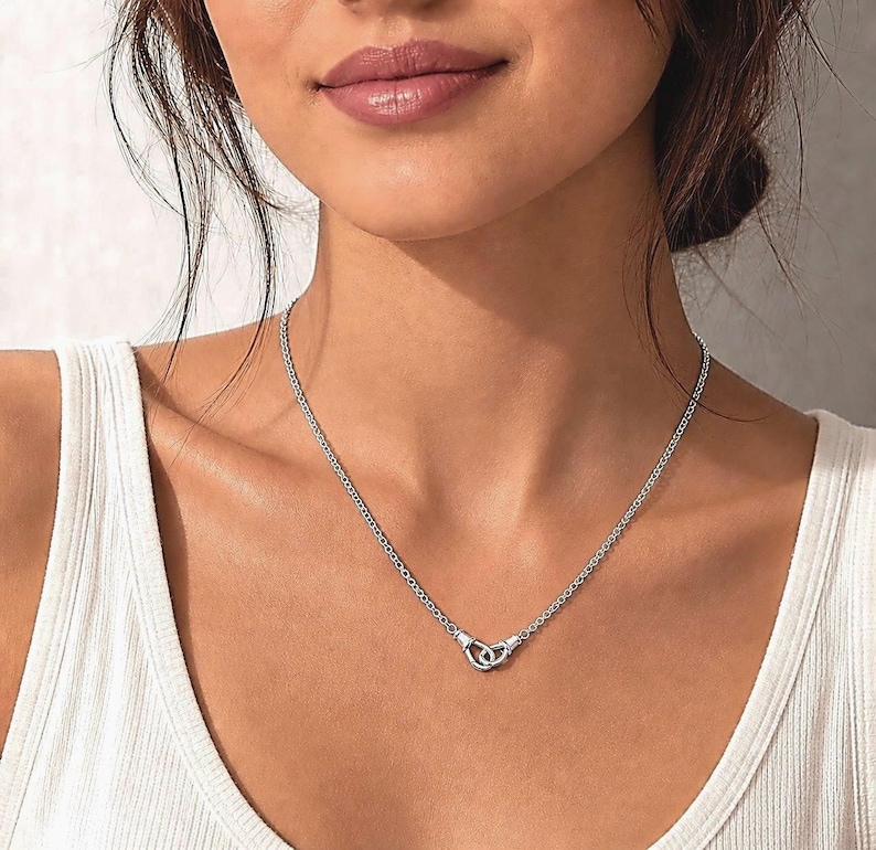 Puede incluir: Un collar plateado con una cadena delicada y un colgante con forma de dos c&iacute;rculos entrelazados. El collar se lleva alrededor del cuello, y el colgante est&aacute; centrado en el pecho. El fondo es de color neutro.