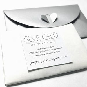 Puede incluir: Primer plano de un sobre plateado con un recorte en forma de coraz&oacute;n y un papel blanco con el texto "SLVR+GLD JEWELRY CO" y detalles del producto. Tambi&eacute;n es visible el texto "prepare for compliments!".