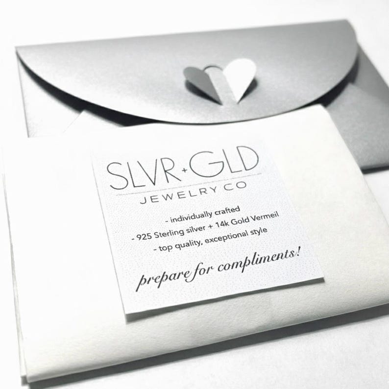 Puede incluir: Primer plano de un sobre plateado con un recorte en forma de coraz&oacute;n y un papel blanco con el texto "SLVR+GLD JEWELRY CO" y detalles del producto. Tambi&eacute;n es visible el texto "prepare for compliments!".