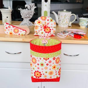 May include: A fabric sewing organizer with a floral pattern in red, yellow, and green. A matching pincushion and small zippered pouch are on a wooden surface. A teapot and teacup are also visible.