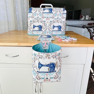 May include: A sewing machine cover, needle case, and a hanging organizer with a matching floral and sewing machine pattern in blue, pink, and white. The organizer has a light blue interior and holds pink scissors.