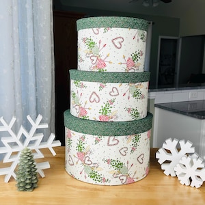 May include: Three round decorative storage boxes stacked on a wooden table. The boxes are white with a repeating pattern of hearts, arrows, and floral elements. Each box has a green patterned rim. Decorative white snowflakes and a green pine tree candle are also on the table.