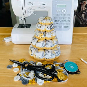 May include: A stack of fabric pumpkins with a floral pattern sits on a wooden table, next to sewing supplies. A white sewing machine is in the background. Scissors, buttons, and a measuring tape are scattered around the pumpkins.
