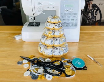 Op de afbeelding: Een stapel stoffen pompoenen met een bloemenpatroon op een houten tafel, naast naaimaterialen. Een witte naaimachine staat op de achtergrond. Schaar, knopen en een meetlint liggen rond de pompoenen.