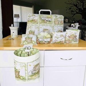 May include: A collection of decorative kitchen items with a green apple theme. Includes a large cutting board, pillows, a canister, and a hanging container. The items feature illustrations of apples, baskets, and farm scenes. The color palette is primarily green and white.