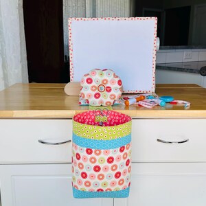 May include: A fabric sewing organizer featuring a hanging cylindrical pouch with a floral pattern in red, green, and light blue, topped with a matching round pin cushion. A white rectangular board with a red floral border stands behind the pouch.