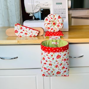 May include: A sewing kit with a floral pattern. The kit includes a pincushion, a needle holder, and a hanging basket. The pincushion is round and has a button in the center. The needle holder is a wooden board with a fabric cover. The basket has a red trim.