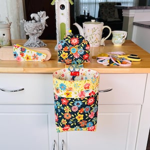 May include: A floral print scissor holder with a matching pin cushion and other sewing accessories. The holder is a cylinder shape with a yellow and navy blue floral pattern. A pair of scissors is inside the holder.