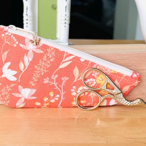 Op de afbeelding: Een koraalkleurig stoffen etui met rits, voorzien van een wit bloemenpatroon en een kleine gouden bijenbedel aan de ritssluiting. Voor het etui ligt een goudkleurige borduurschaar in de vorm van een ooievaar met gedetailleerde handvatten.