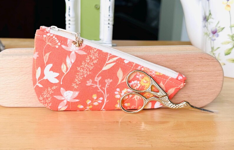 Op de afbeelding: Een koraalkleurig stoffen etui met rits, voorzien van een wit bloemenpatroon en een kleine gouden bijenbedel aan de ritssluiting. Voor het etui ligt een goudkleurige borduurschaar in de vorm van een ooievaar met gedetailleerde handvatten.
