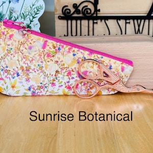 Op de afbeelding: Een driehoekig stoffen etui met een kleurrijk bloemenpatroon in geel, roze en blauw met een felroze rits. Een ros&eacute;gouden borduurschaar in de vorm van een ooievaar met een roze punt ligt voor het etui. De tekst luidt Sunrise Botanical.