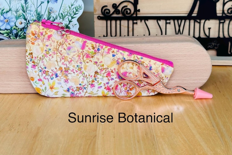 Op de afbeelding: Een driehoekig stoffen etui met een kleurrijk bloemenpatroon in geel, roze en blauw met een felroze rits. Een ros&eacute;gouden borduurschaar in de vorm van een ooievaar met een roze punt ligt voor het etui. De tekst luidt Sunrise Botanical.