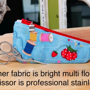Op de afbeelding: Een lichtblauw stoffen etui met een rode rits en een zilverkleurige bedel in de vorm van een garenklosje. Het etui heeft een print van garenklosjes, aardbeien en een ruitpatroon. Een roestvrijstalen schaar ligt voor het etui.