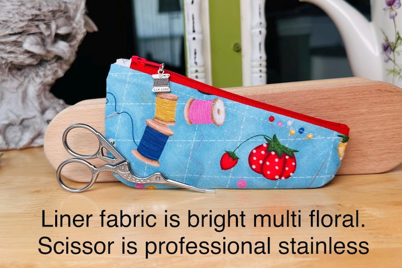 Op de afbeelding: Een lichtblauw stoffen etui met een rode rits en een zilverkleurige bedel in de vorm van een garenklosje. Het etui heeft een print van garenklosjes, aardbeien en een ruitpatroon. Een roestvrijstalen schaar ligt voor het etui.