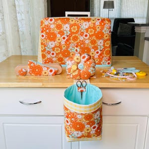 May include: A sewing machine cover, pin cushion, and other sewing accessories are displayed on a wooden table. The items feature a retro orange floral pattern. A hanging fabric basket holds scissors.