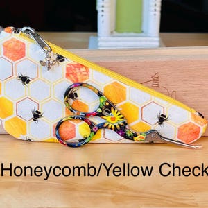 Op de afbeelding: Een driehoekig stoffen etui met een honingraat- en bijenpatroon, voorzien van een gele rits en een kleine zilveren bijenbedel. Een meerkleurige schaar met bloemenpatroon ligt op het etui. De tekst luidt Honeycomb/Yellow Check.