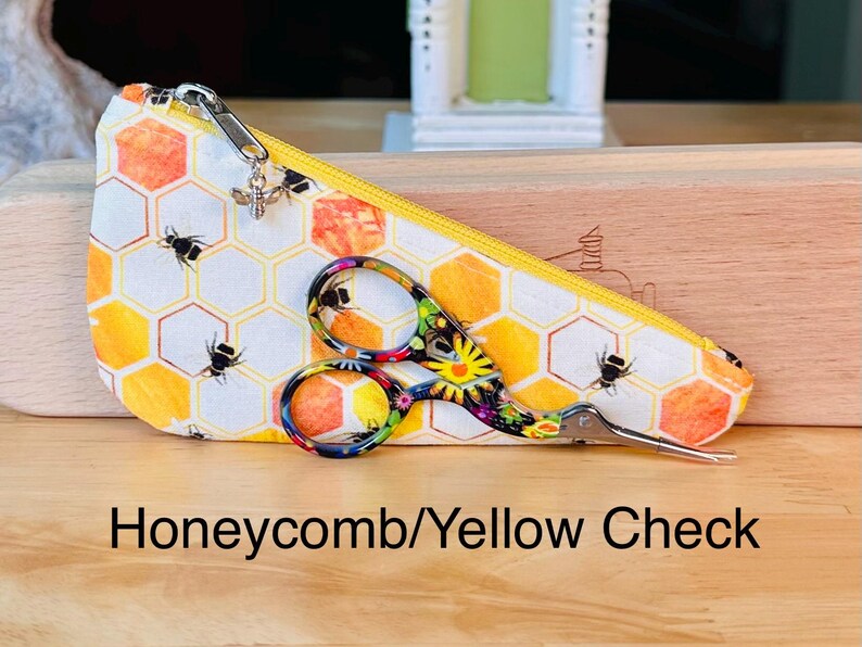 Op de afbeelding: Een driehoekig stoffen etui met een honingraat- en bijenpatroon, voorzien van een gele rits en een kleine zilveren bijenbedel. Een meerkleurige schaar met bloemenpatroon ligt op het etui. De tekst luidt Honeycomb/Yellow Check.