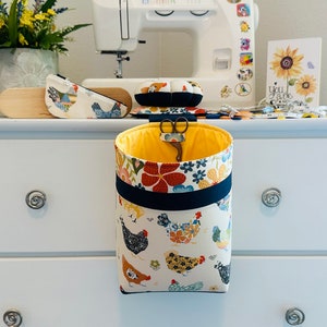May include: A fabric hanging organizer with a yellow interior and a chicken pattern. The organizer is hanging from a white dresser. A pair of scissors is inside the organizer. Other sewing accessories are on the dresser.