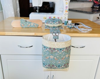 May include: A fabric sewing organizer set featuring a hanging waste bin, a matching pin cushion, and a wooden tailor's clapper, all covered in a teal and beige floral bird print fabric. The set includes a pair of metal scissors inside the bin.