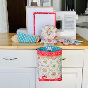 May include: A fabric sewing organizer set featuring a hanging waste bin with a floral pattern, a matching round pin cushion with a central button, a rectangular pressing board, and a small teal patterned fabric piece, all arranged near a sewing machine.