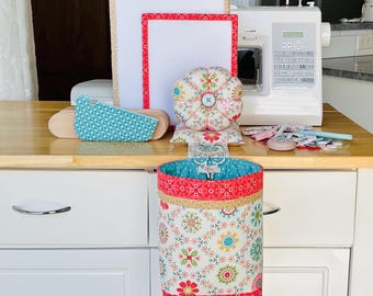 May include: A fabric sewing organizer set featuring a hanging waste bin with a floral pattern, a matching round pin cushion with a central button, a rectangular pressing board, and a small teal patterned fabric piece, all arranged near a sewing machine.
