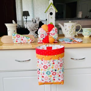 May include: A white and floral fabric bin with red and orange trim hangs from a white cabinet. Other items on the wooden countertop include a pin cushion, a birdhouse, a teapot, and a teacup. The items have a floral theme.