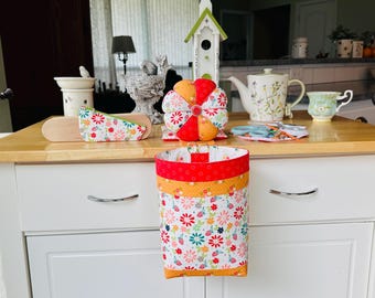 May include: A white and floral fabric bin with red and orange trim hangs from a white cabinet. Other items on the wooden countertop include a pin cushion, a birdhouse, a teapot, and a teacup. The items have a floral theme.