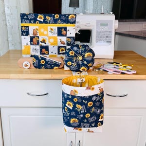 May include: A sewing machine with a navy blue and yellow fabric cover, a pin cushion, a needle case, and a hanging basket. The fabric features sunflowers, bees, and the text "Bee Kind and Free".