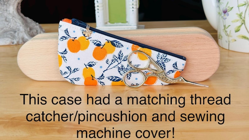 Op de afbeelding: Een wit stoffen etui met rits en een patroon van sinaasappels en blauwe bladeren, versierd met een kleine gouden bijenbedel aan de ritssluiting. Een zilveren ooievaarsschaartje ligt op het etui, dat voor een houten kistje is geplaatst.