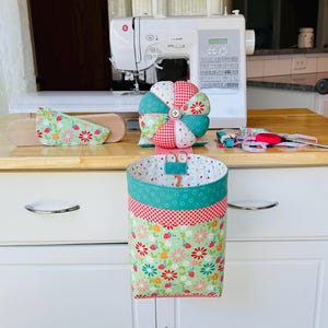 May include: A sewing-themed image featuring a white sewing machine, a fabric basket, and other sewing accessories. The basket has a floral pattern with red, green, and white accents. A pincushion and a wooden cutting board with fabric are also visible.