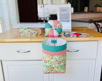 May include: A fabric sewing organizer with a hanging pocket featuring a green floral and strawberry print, a red and white gingham trim, and a teal band. It includes a matching round, multi-sectioned pin cushion with a central button detail.
