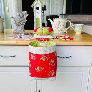 May include: A red fabric waste basket with a floral pattern, hanging from a white cabinet. The basket's interior is green. A pincushion, scissors, and other sewing notions are on the wooden countertop.