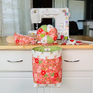 May include: A sewing-themed still life featuring a white sewing machine, a pincushion, a fabric basket, and other sewing notions. The fabric items have a floral pattern in shades of coral, green, and white. A wooden pressing ham and spools of thread are also visible.