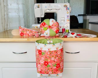 May include: A fabric hanging storage basket with a coral floral print and white polka dot trim, accompanied by a matching round segmented pincushion, a small zippered pouch, and a sewing machine with various sewing notions and tools.