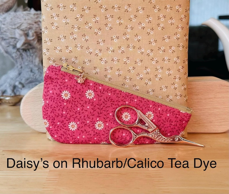 Op de afbeelding: Een roze stoffen etui met een wit madeliefjes- en rankenpatroon, voorzien van een gouden rits met een kleine bedel, geplaatst voor een beige stoffen vierkant met een wit bloemenpatroon. Een versierde goudkleurige metalen schaar ligt op het etui.