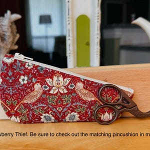 Op de afbeelding: Een rood stoffen etui met een witte rits en een herhalend patroon van vogels en bloemen, vergezeld door een donker metalen schaar met een vogelvormig handvat. De tekst luidt: Strawberry Thief. Be sure to check out the matching pincushion in my shop.