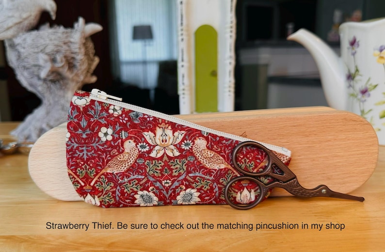 Op de afbeelding: Een rood stoffen etui met een witte rits en een herhalend patroon van vogels en bloemen, vergezeld door een donker metalen schaar met een vogelvormig handvat. De tekst luidt: Strawberry Thief. Be sure to check out the matching pincushion in my shop.