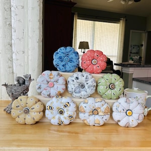 May include: Nine fabric pincushions shaped like flowers in various colors and patterns, including blue, red, green, white, and yellow. The pincushions are arranged in a pyramid shape on a wooden table.