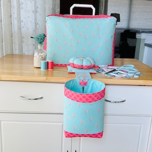 May include: A sewing kit set with a flamingo pattern on a light blue background and pink trim. The set includes a sewing machine cover, a pin cushion, a scissors holder, and other sewing accessories. The items are displayed on a white and wood-tone cart.