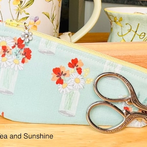 Op de afbeelding: Een lichtgroen stoffen etui met ritssluiting, voorzien van een herhalend patroon van bloemen in glazen potten, een gele rits en een kleine zilveren bloembedel. Een decoratieve zilverkleurige metalen borduurschaar ligt op het etui.