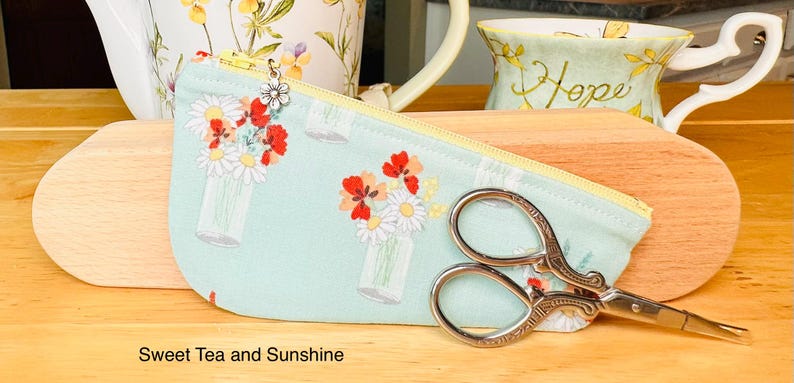 Op de afbeelding: Een lichtgroen stoffen etui met ritssluiting, voorzien van een herhalend patroon van bloemen in glazen potten, een gele rits en een kleine zilveren bloembedel. Een decoratieve zilverkleurige metalen borduurschaar ligt op het etui.