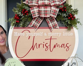 Könnte beinhalten: Ein runder Weihnachtsschmuck mit dem Text "Have yourself a merry little Christmas" in roter Schrift. Das Schild hat einen Holzmaserungshintergrund, einen roten und schwarzen Rand und ist mit Grün und einer karierten Schleife verziert.
