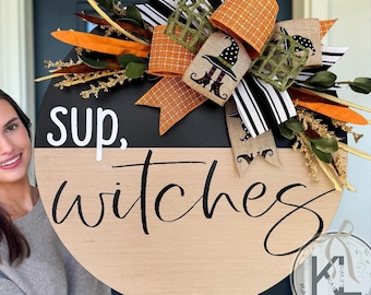 May include: A round wooden door sign with a black top half and a natural wood bottom half. The text "sup, witches" is printed in white and black. It is decorated with a large bow featuring orange, black and white stripes, and burlap ribbons with a witch hat and boot print, plus artificial greenery and orange accents.