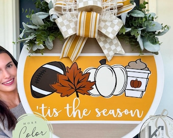 May include: A round wooden door sign featuring a yellow background with a football, a brown maple leaf, a white pumpkin, and a coffee cup graphic. The text reads 'tis the season. It is topped with a large bow in yellow and white stripes and checks.