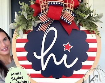 May include: A round wooden door sign featuring horizontal red and white stripes and a central navy blue scalloped circle with the white text hi and a small red star. The sign is decorated with green foliage and a large red, white, and blue bow.