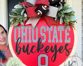 May include: A round wooden door sign featuring a red background with the gray text OHIO STATE and the black script text Buckeyes. A large gray block letter O is centered at the bottom. A decorative bow with football patterns sits on top.