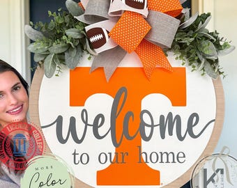 Puede incluir: Un letrero de puerta redondo de madera con una letra T grande de color naranja, el texto "welcome to our home" en gris y un lazo decorativo con lunares naranjas, tela gris y gráficos de fútbol americano, con follaje artificial verde.