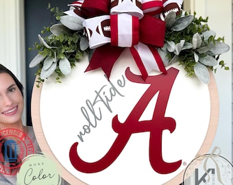 Op de afbeelding: Een rond houten deurbord met een witte achtergrond, een grote rode letter A en de handgeschreven tekst roll tide. Het bord is versierd met een grote strik met football-afbeeldingen en rood-witte strepen, geaccentueerd door groen gebladerte.