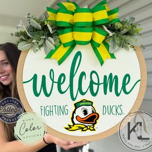 May include: A round wooden welcome sign with a white center and a light wood border. The sign features the words "welcome" and "Fighting Ducks" in green script, along with the Oregon Ducks logo. It is decorated with a green and yellow bow and greenery.