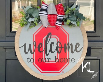 May include: A round wooden door hanger with a gray background and a red "O" with a black outline. The text "welcome to our home" is written in black cursive script. A red and white bow with green foliage is attached to the top of the hanger.